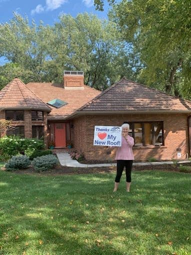 Homeowner holding an Innovative Roofing thank-you sign in front of a brick home with a new roof