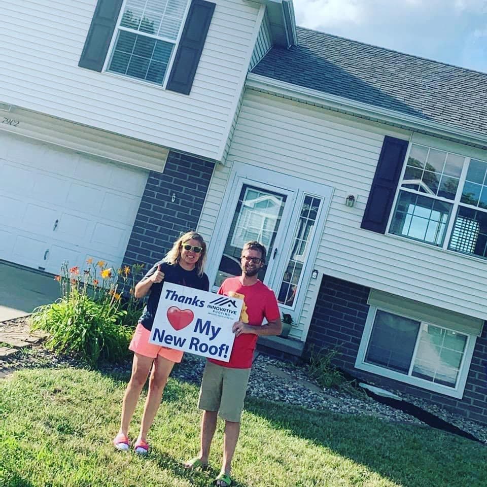 Couple giving a thumbs-up while holding a Thanks My New Roof Innovative Roofing sign on their front lawn