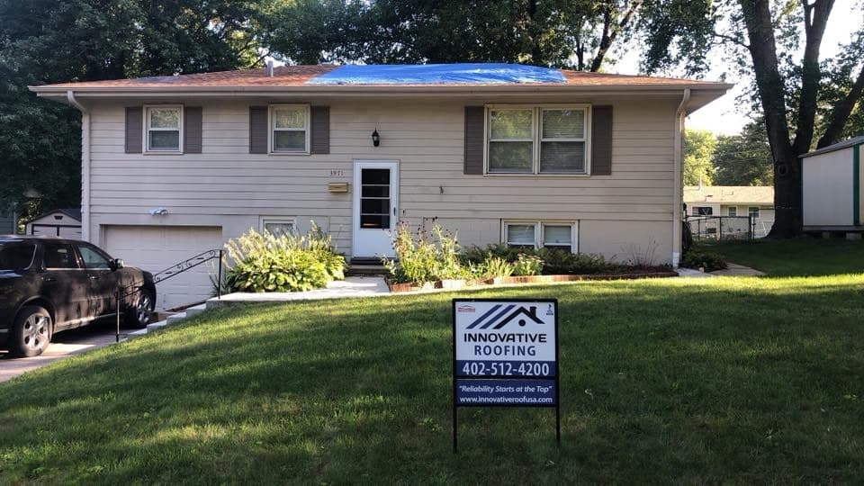 Split-level home with a blue emergency tarp on the roof and an Innovative Roofing yard sign in the lawn