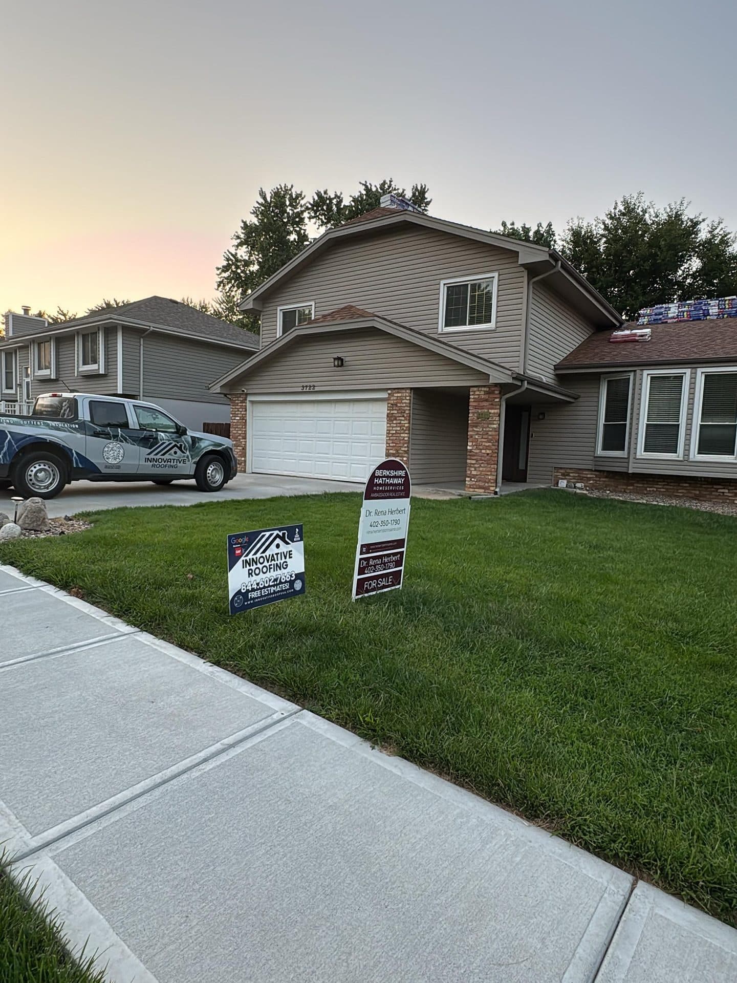 Tan two-story home at dusk with Innovative Roofing branded truck in the driveway and shingle bundles on the roof
