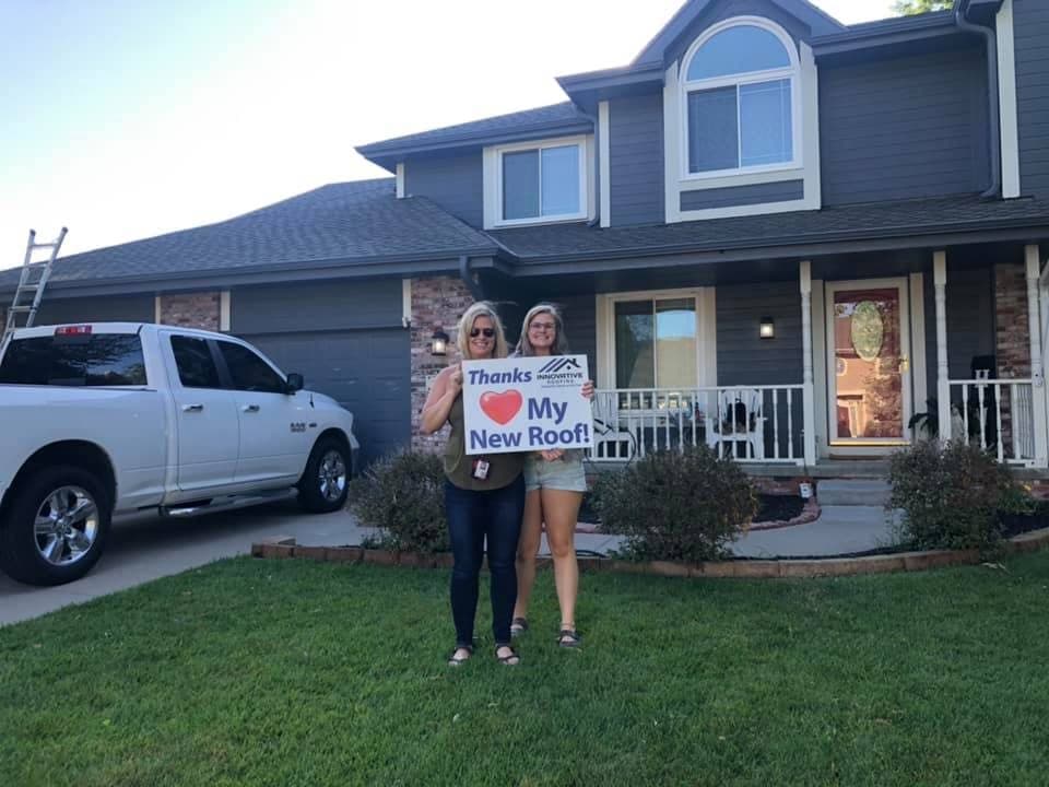 Two homeowners holding a Thanks My New Roof sign in front of a two-story gray home