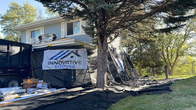 Light blue home mid roof replacement with Innovative Roofing banner on debris netting and materials staged on the lawn